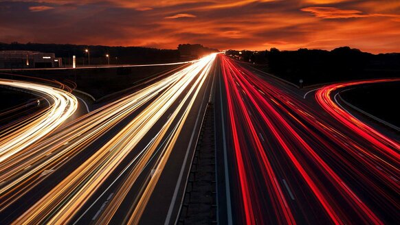 Langzeitbelichtung: Rote und weiße Streifen von Autolichtern auf einer Autobahn vor orangefarbenem Himmel.